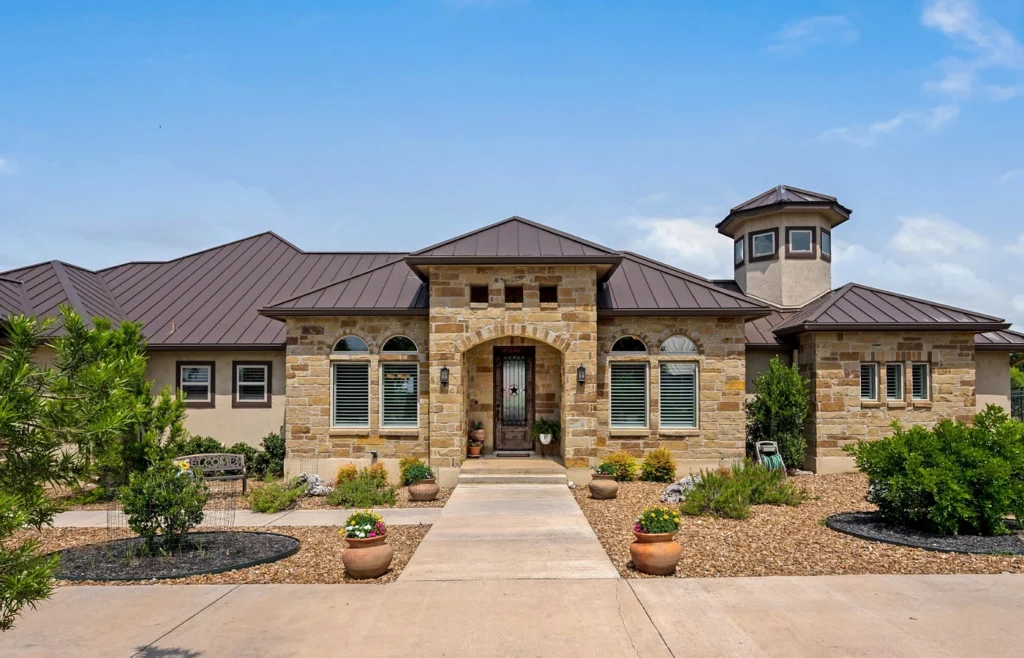 Single-story limestone home with a standing-seam metal roof, an arched front entry, a turret and a stone-mulch front yard under a blue Texas sky, representing buying or selling a home in Whitney.