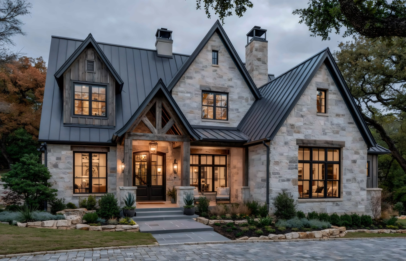 Two-story limestone home with a metal roof, timber entry and live oaks at dusk, representing homes for sale in Central Texas