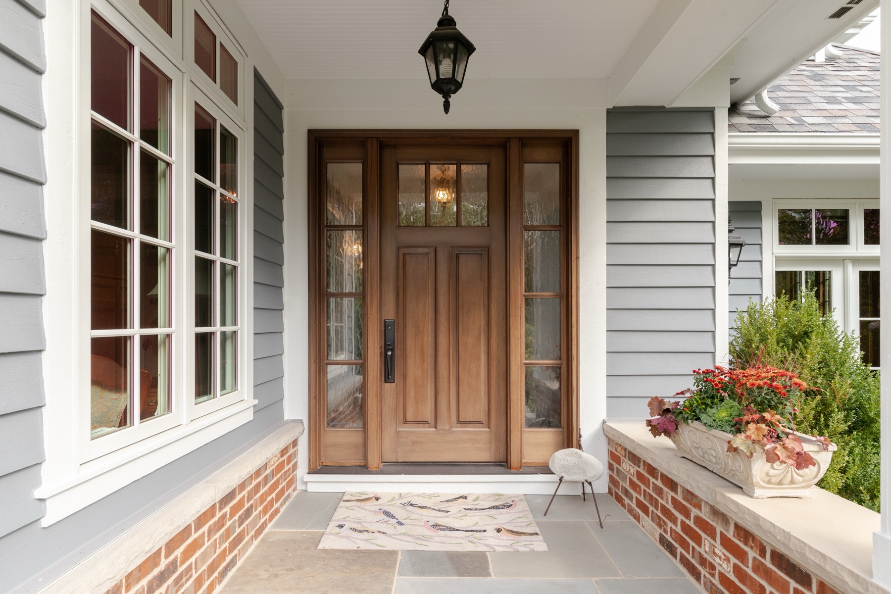 Covered front porch with a wood entry door, glass sidelights, a lantern and a planter of fall flowers on a brick ledge