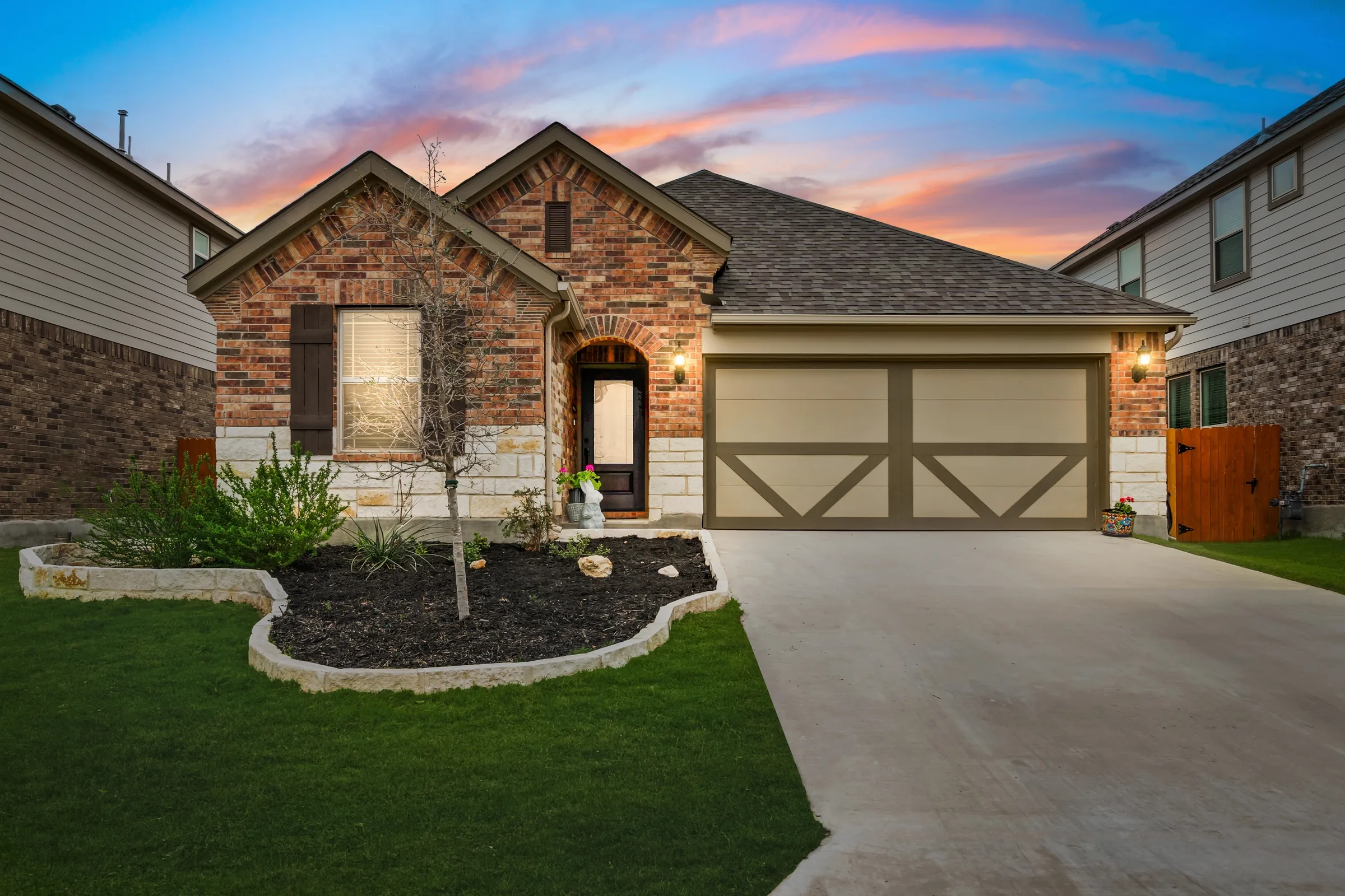A single-story brick home with a two-car garage and a landscaped bed under a pink sunset sky