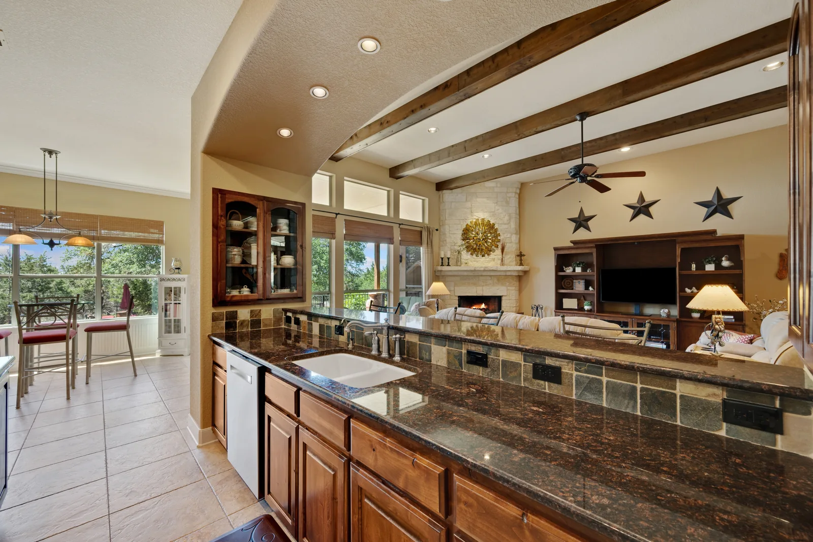 An open kitchen with granite counters and wood cabinets, a beamed ceiling and a stone corner fireplace