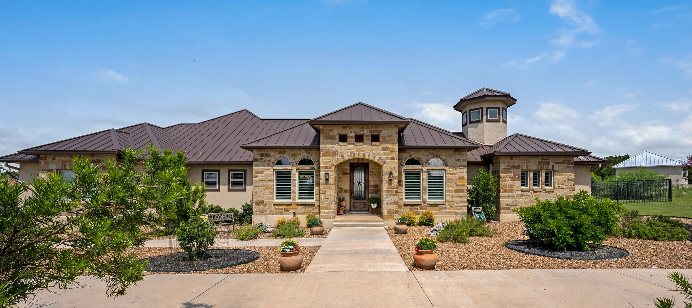 A stone single-story home with a standing-seam metal roof and a corner tower, a xeriscaped yard and a blue sky