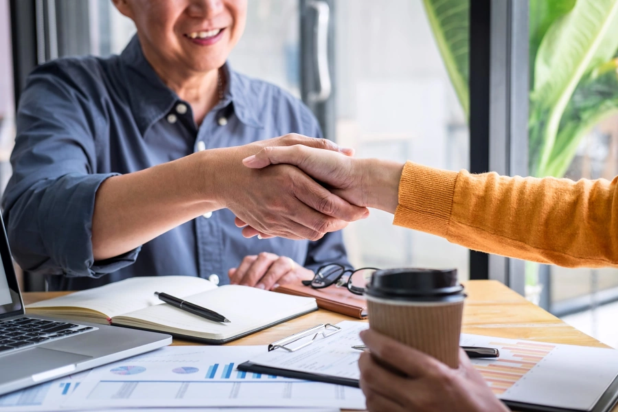 Two people shaking hands across a desk with documents and coffee, representing closing day when selling a home in Whitney